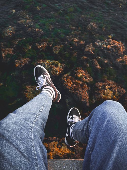 Free Casual view from above of legs in sneakers over vibrant water with rocks below. Stock Photo