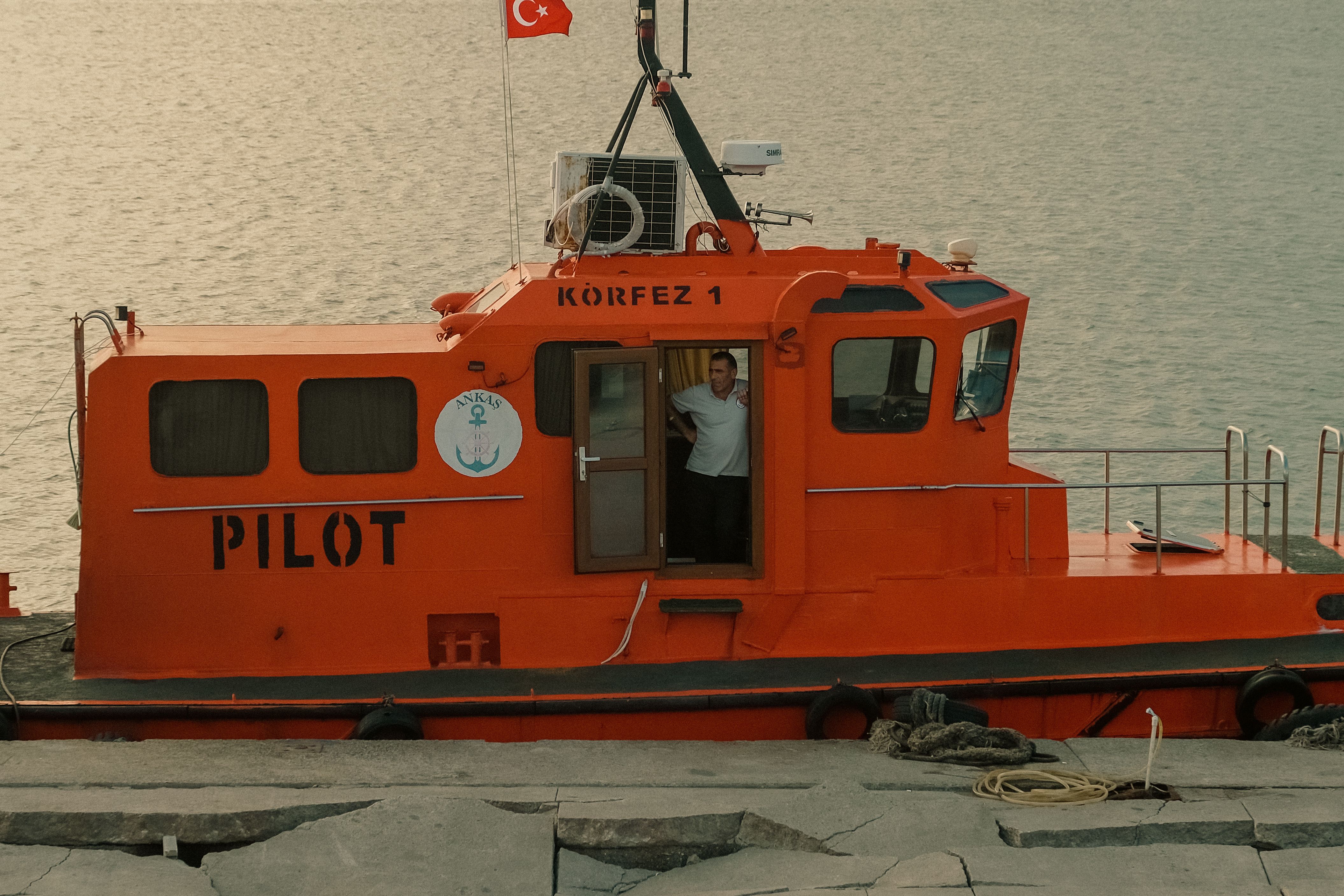 Orange pilot boat moored at harbor with Turkish flag, featuring a man standing in the doorway.