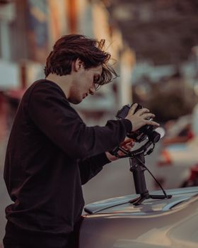 A young man in casual wear adjusts a camera on a car outdoors, capturing urban scenery.