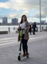 Full body of young thrilled female riding modern scooter on urban street with people under cloudy sky on blurred background