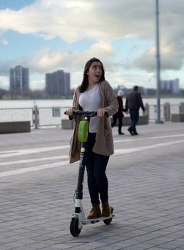 Full body of young thrilled female riding modern scooter on urban street with people under cloudy sky on blurred background