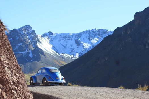 A classic blue Volkswagen Beetle drives along a scenic mountain road with snow-capped peaks.