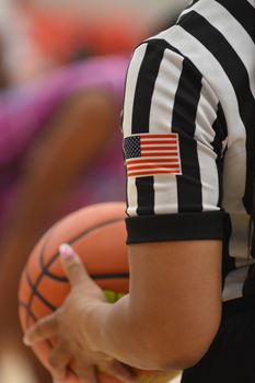 Close-up of a referee holding a basketball with an American flag patch on the sleeve, indoors.