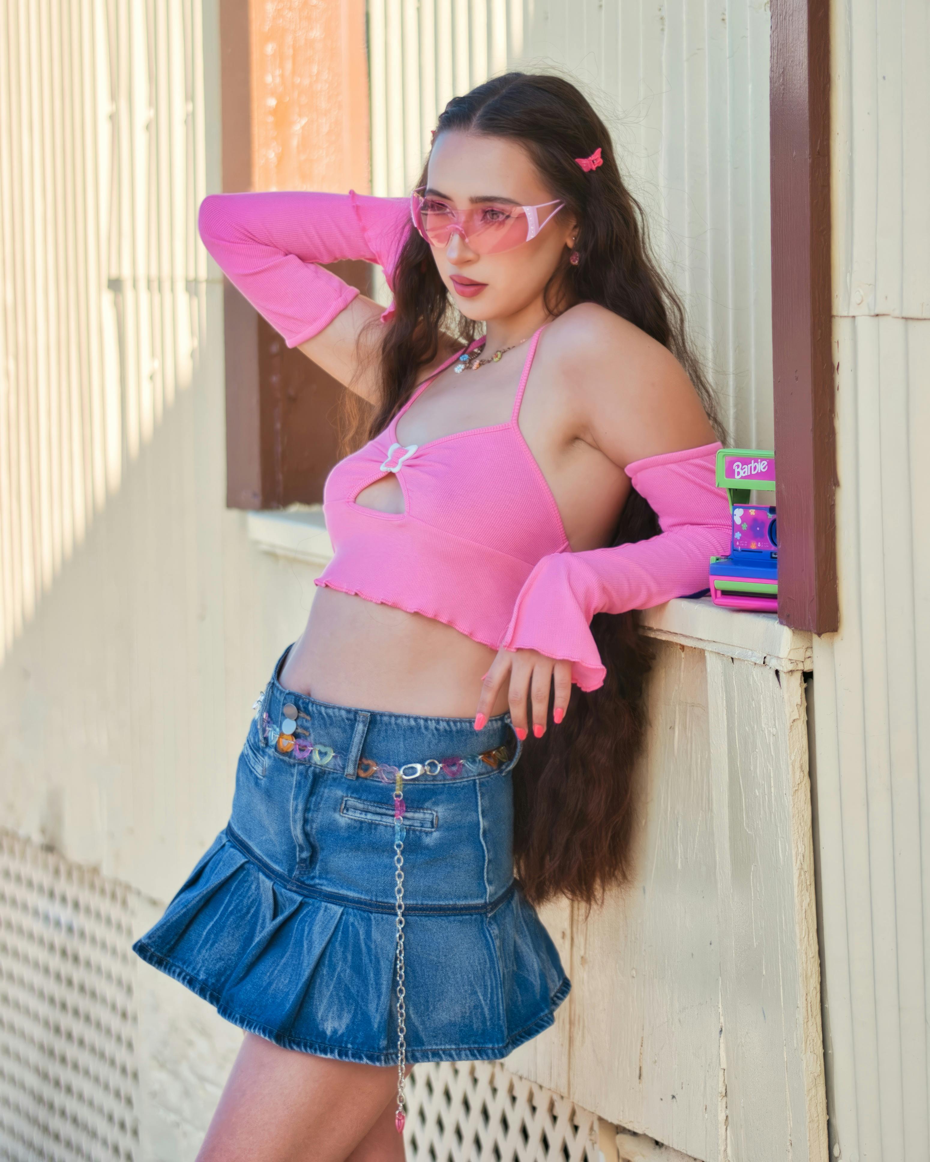 Fashionable woman wearing a pink crop top and denim skirt leaning against a wall, showcasing modern style.