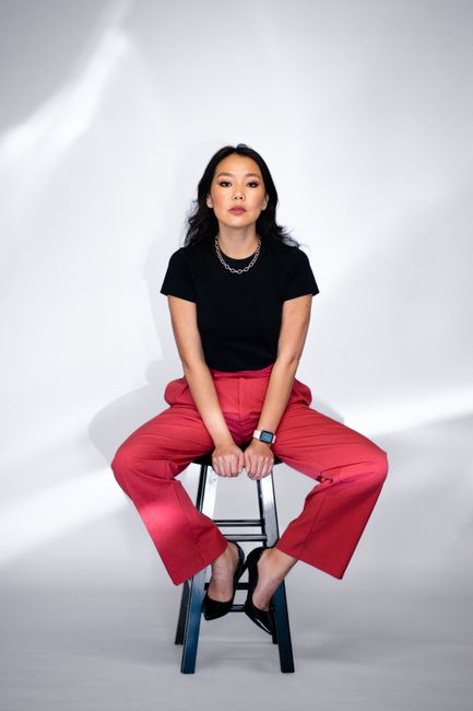 Confident woman in fashionable attire sitting on stool against white background.
