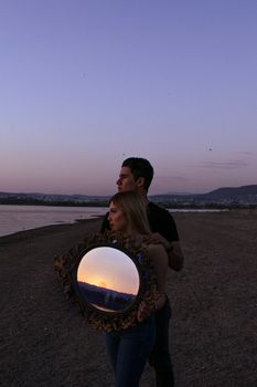 A romantic couple stands by the beach with a mirror reflecting the sunset in León, Mexico.