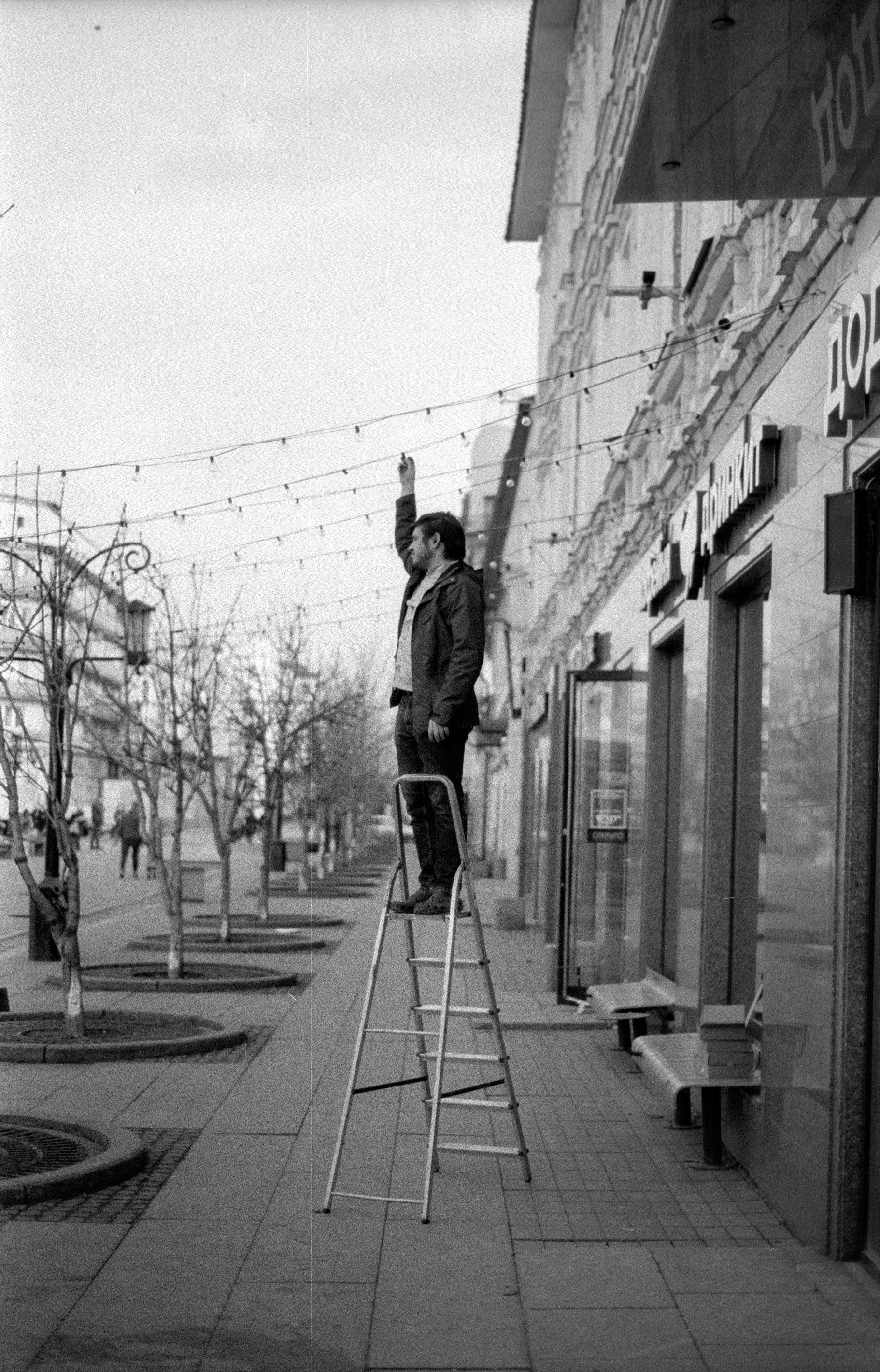 Black and white photo of a man adjusting lights on a ladder in the city.