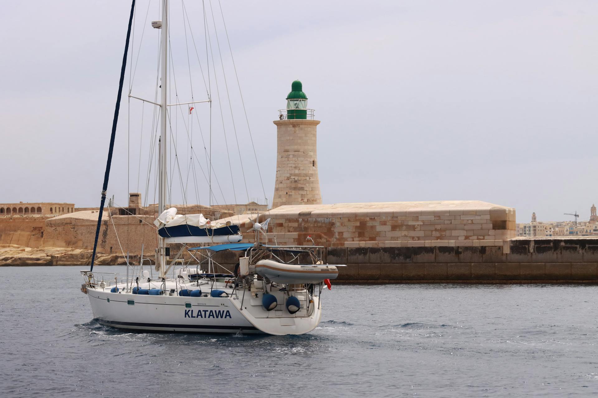A sailboat named Klatawa sails past a historic lighthouse in Valletta, Malta.