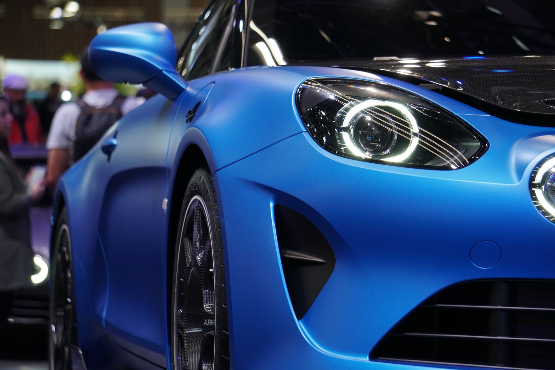 Close-up of a blue concept sports car with illuminated circular headlights on a convention floor, with attendees visible in the background — generic auto show activity, no identifiable venue or signage.