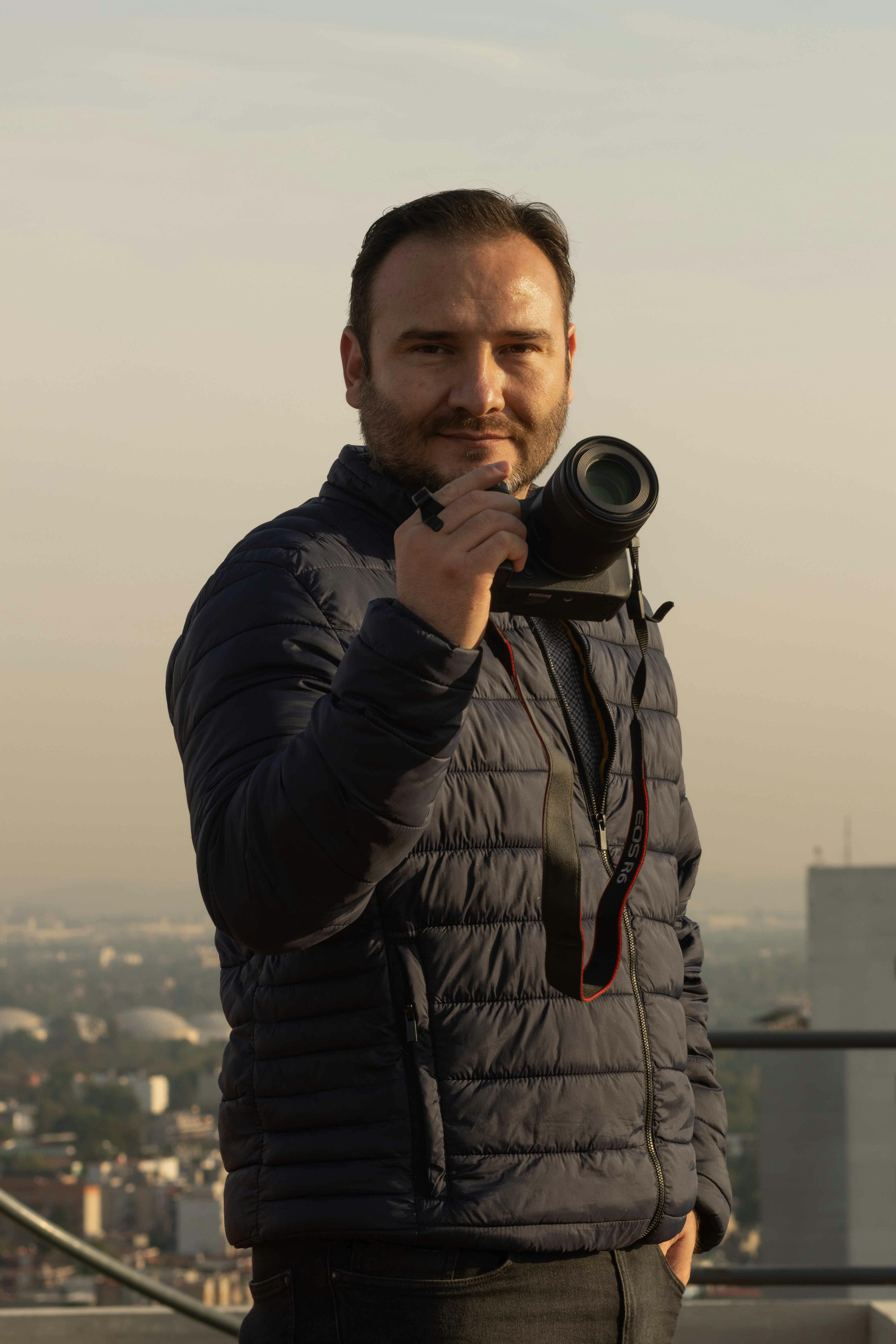 Man in black jacket posing with camera on rooftop during day.