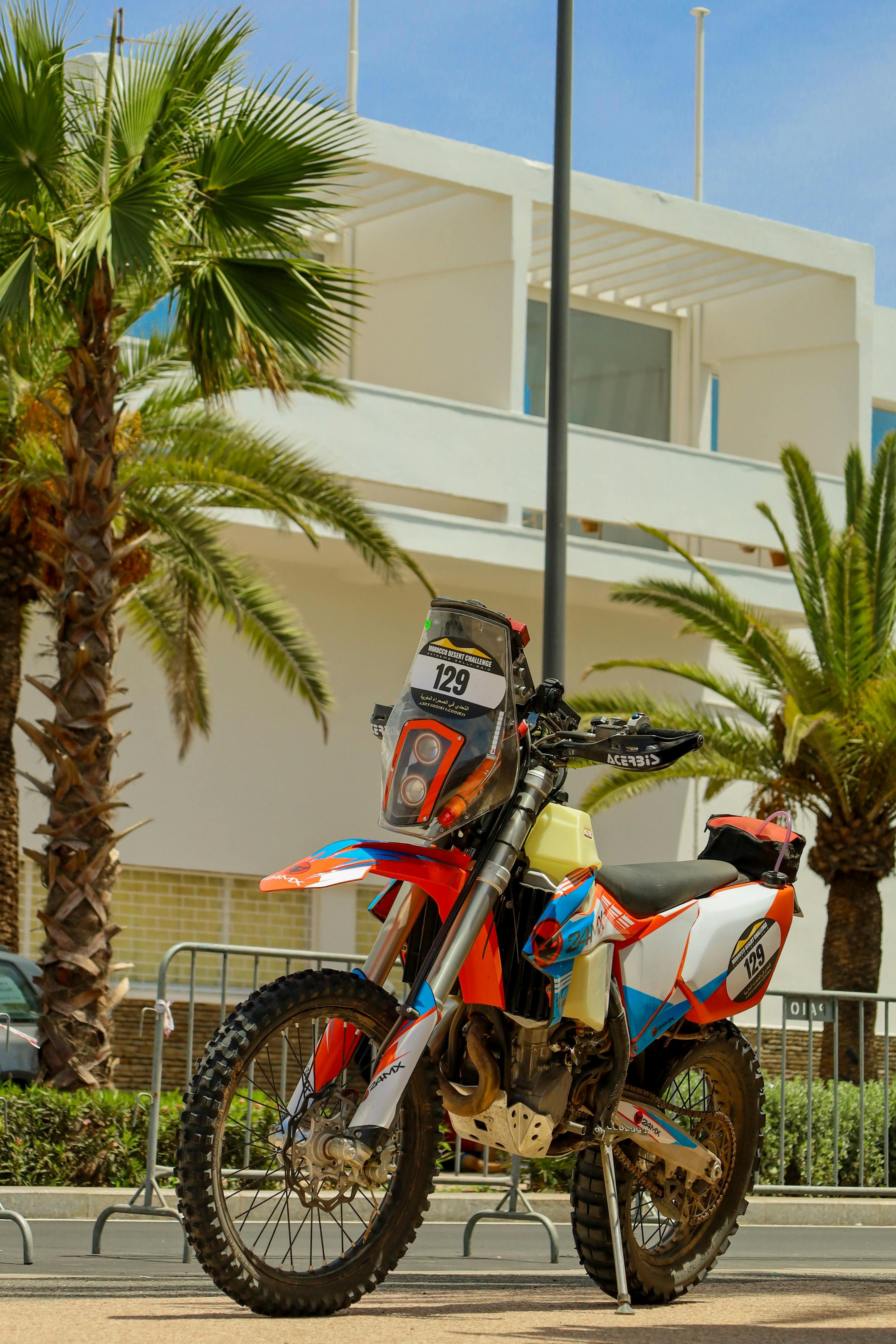 Vibrant motocross bike with number 129 parked amidst palm trees in an urban setting, capturing a tropical adventure vibe.