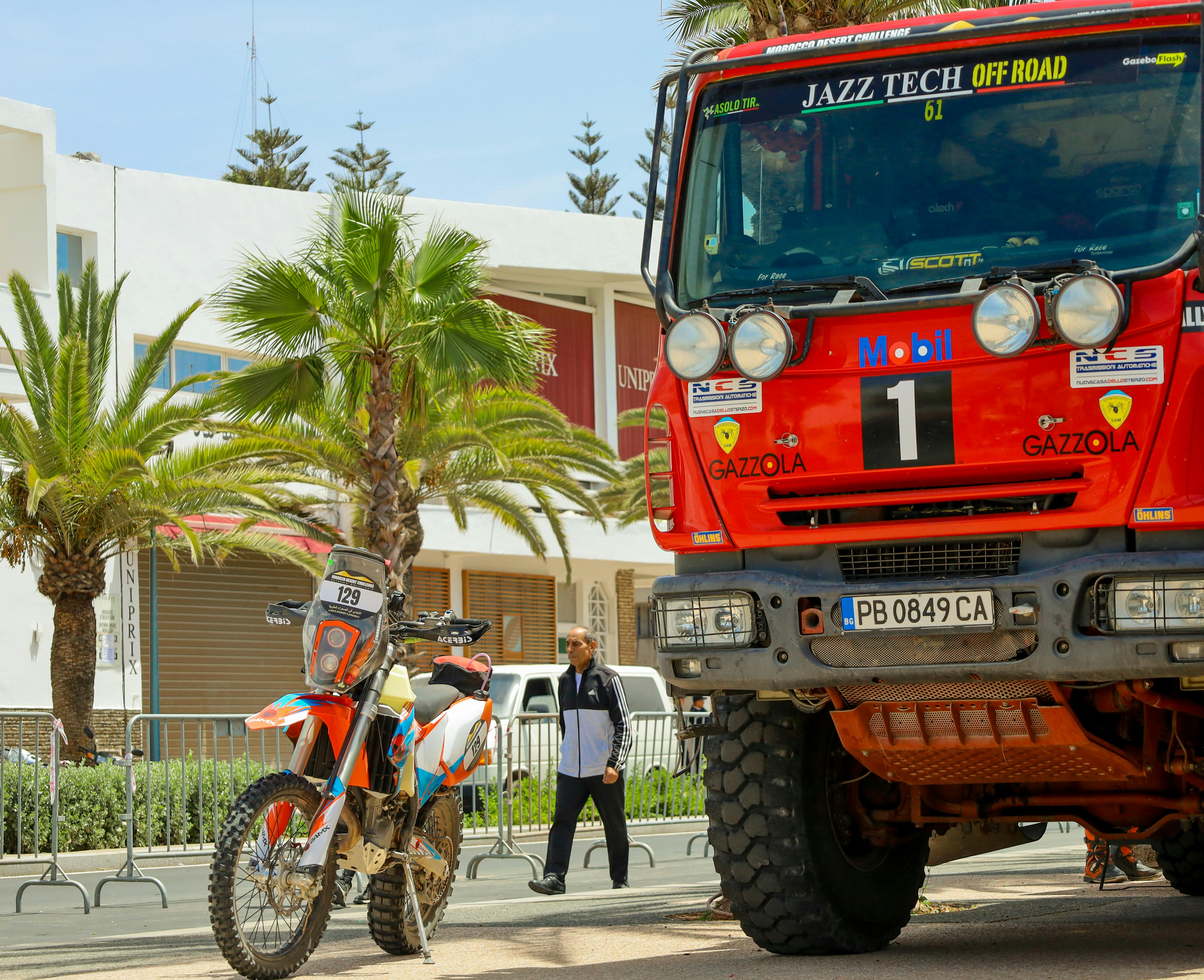 Rally truck and motocross bike displayed outdoors with palm trees.