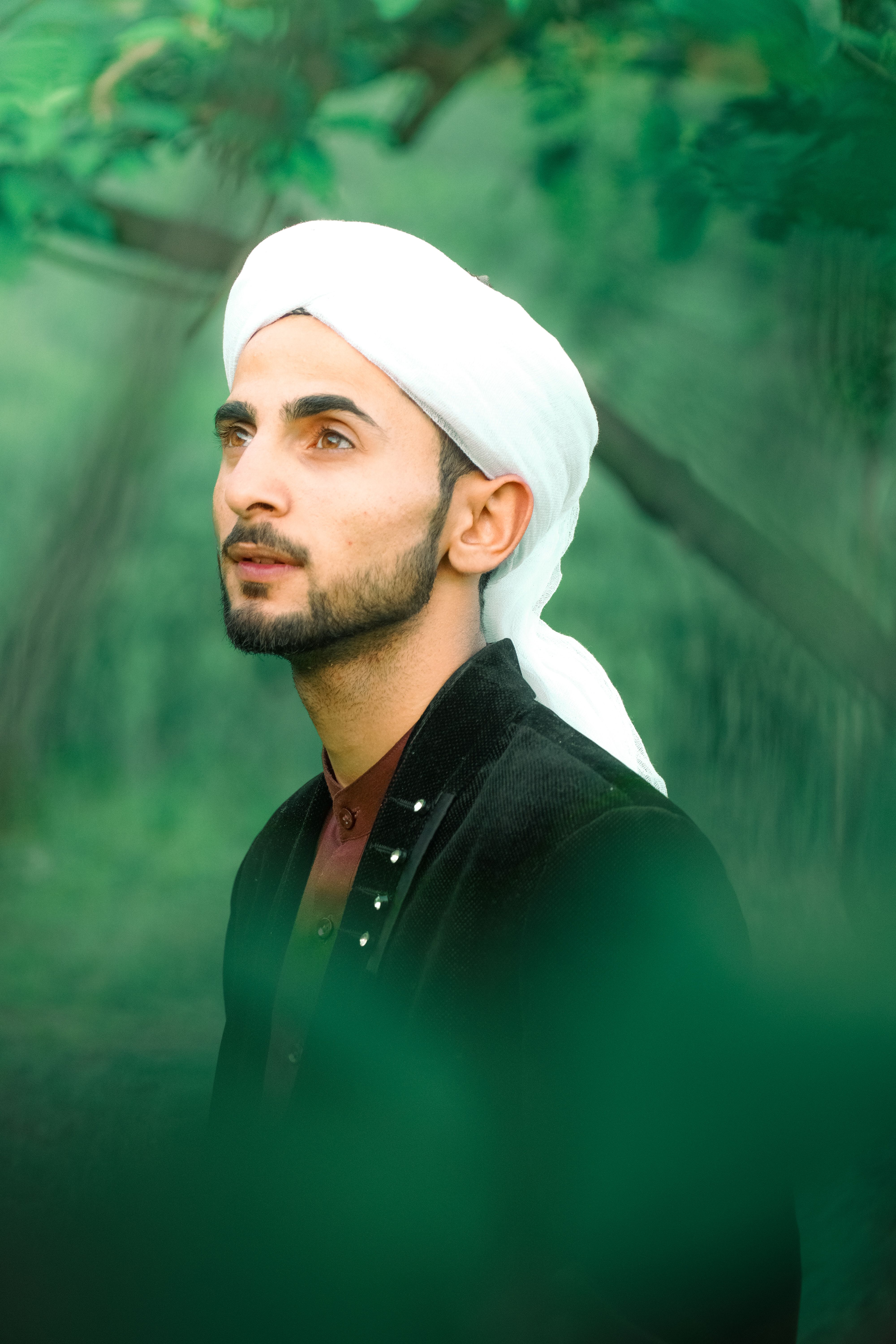 Portrait of a young man wearing a white turban, gazing upwards amidst green foliage.