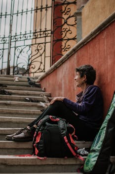 Elderly woman rests on steps with a backpack in a historic town setting, enjoying a moment of reflection.