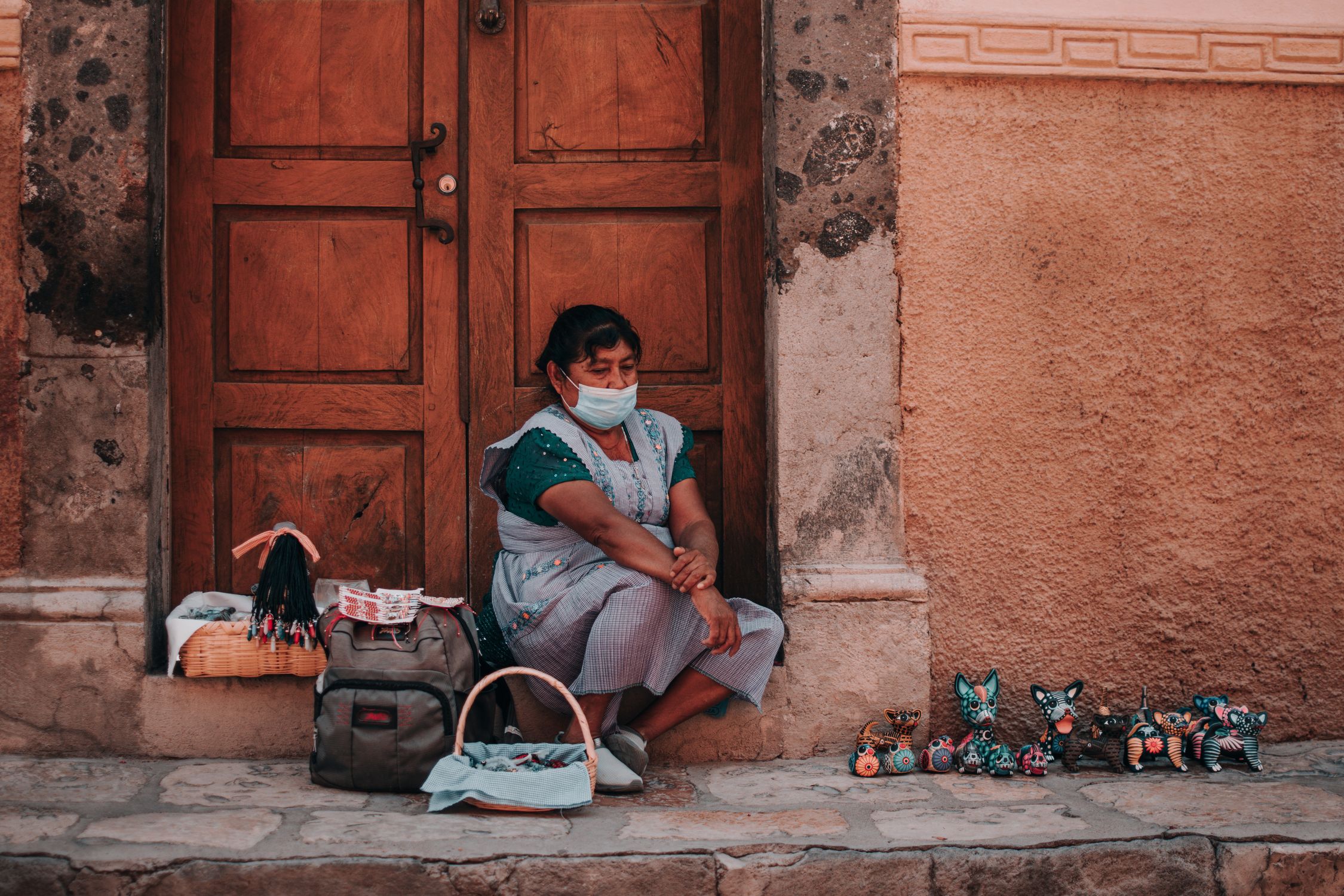 A woman in San Miguel de Allende sits by a wooden door selling handmade crafts while wearing a face mask.