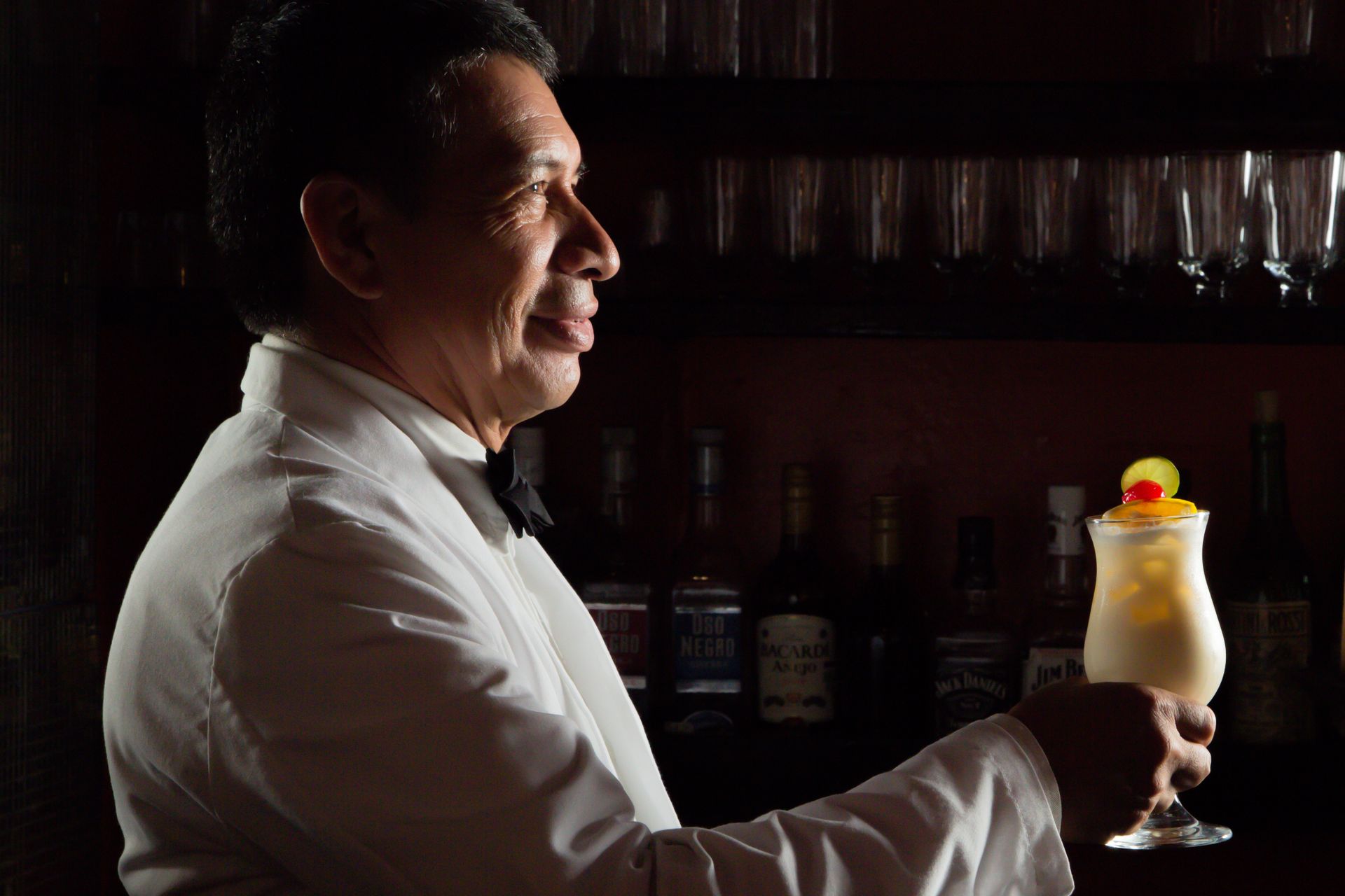 Bartender in a white jacket serves a tropical pina colada cocktail in a dim, cozy bar setting.