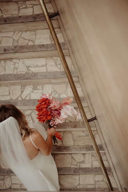 Free Elegant bride in white dress and veil holding a colorful bouquet while walking upstairs. Aerial view. Stock Photo