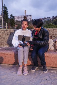 A couple sitting on a sandy bench, operating a drone at the beachfront during daytime.