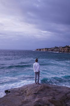 这个人站在岩石海岸上，凝视着多云天空下平静的海面