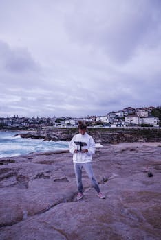 A person flying a drone at a rocky beach with a coastal town in the background under cloudy skies.