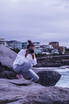 Woman taking a photo on a rocky beach with urban buildings in the background.