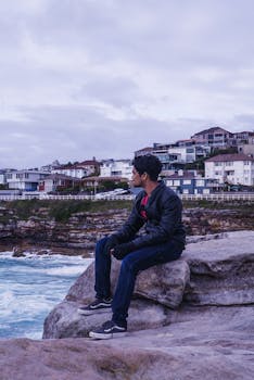 一名男子穿着皮夹克，坐在海边的岩石上，背景是沿海的房屋