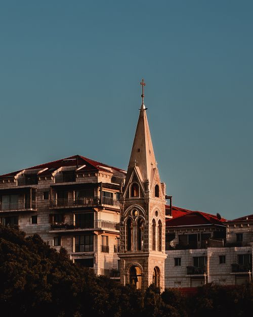 Free A sunlit gothic church tower against a cityscape under a clear sky. Stock Photo