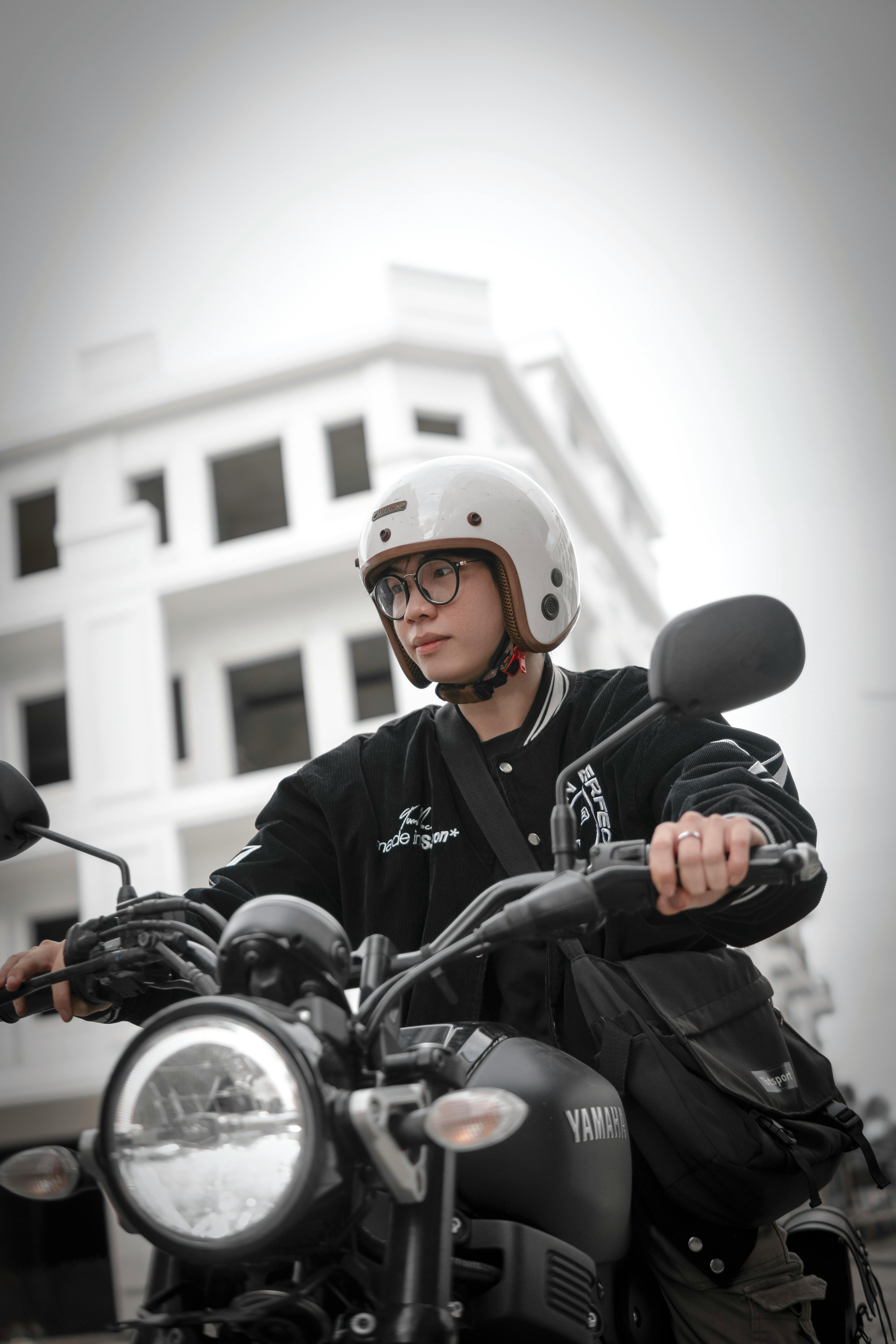 Young adult on a Yamaha motorbike, wearing glasses and helmet, in an urban setting.