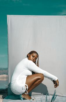A young woman crouches elegantly in a white dress against a minimal outdoor backdrop.