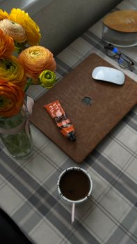 Top view of a workspace featuring flowers, a laptop, cup of coffee, and a tube on a plaid tablecloth.
