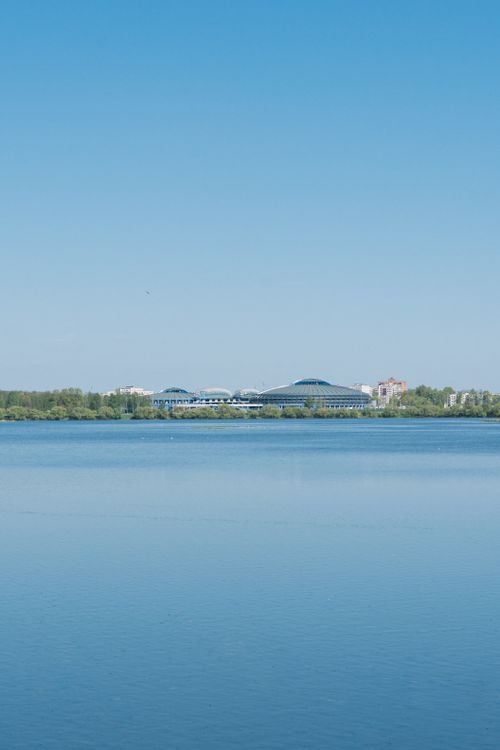 Free Serene lake scene with a sports arena and modern architecture in Minsk, Belarus under a clear blue sky. Stock Photo
