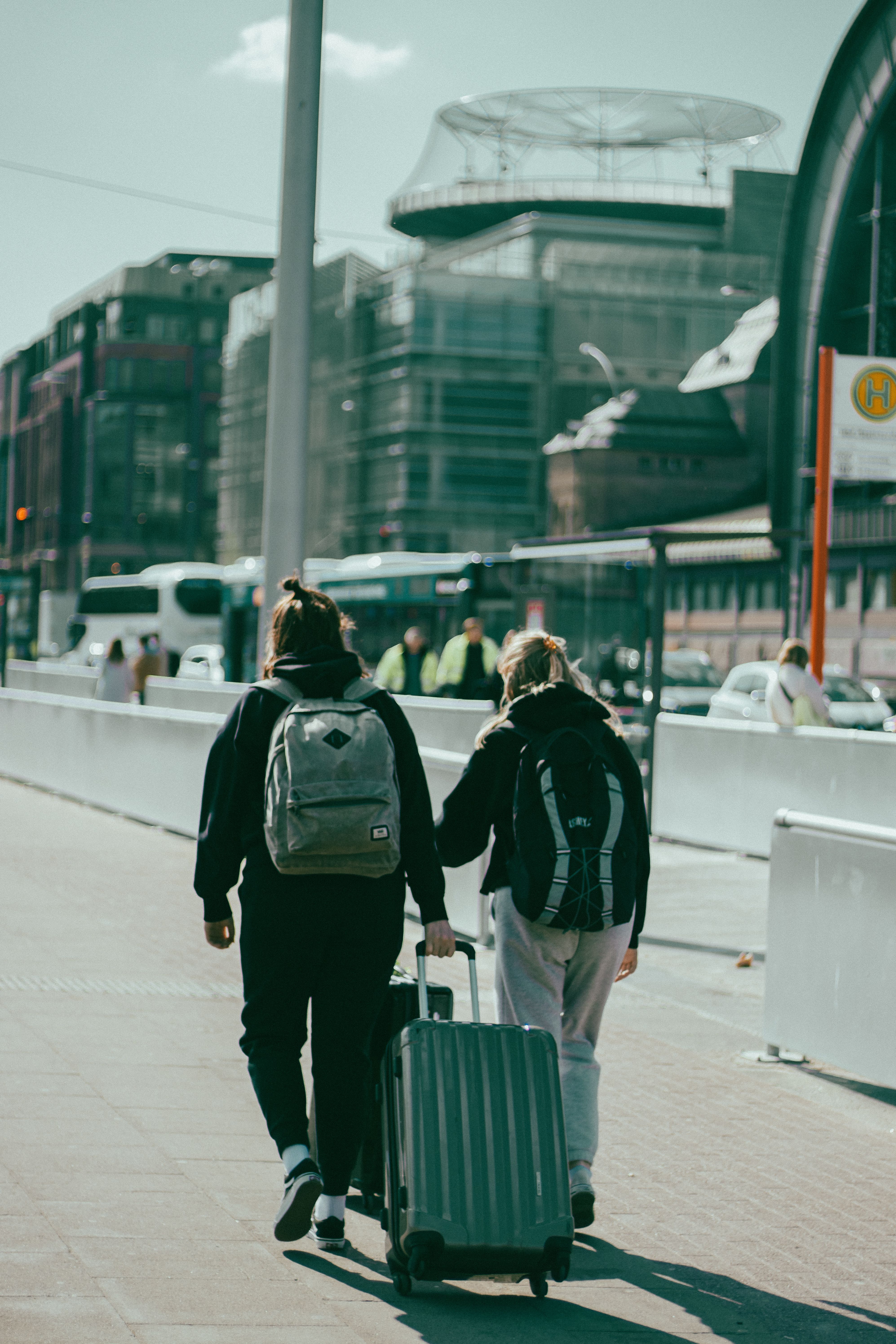 A couple of travelers walk through a sunlit city, dragging their suitcase on the sidewalk.