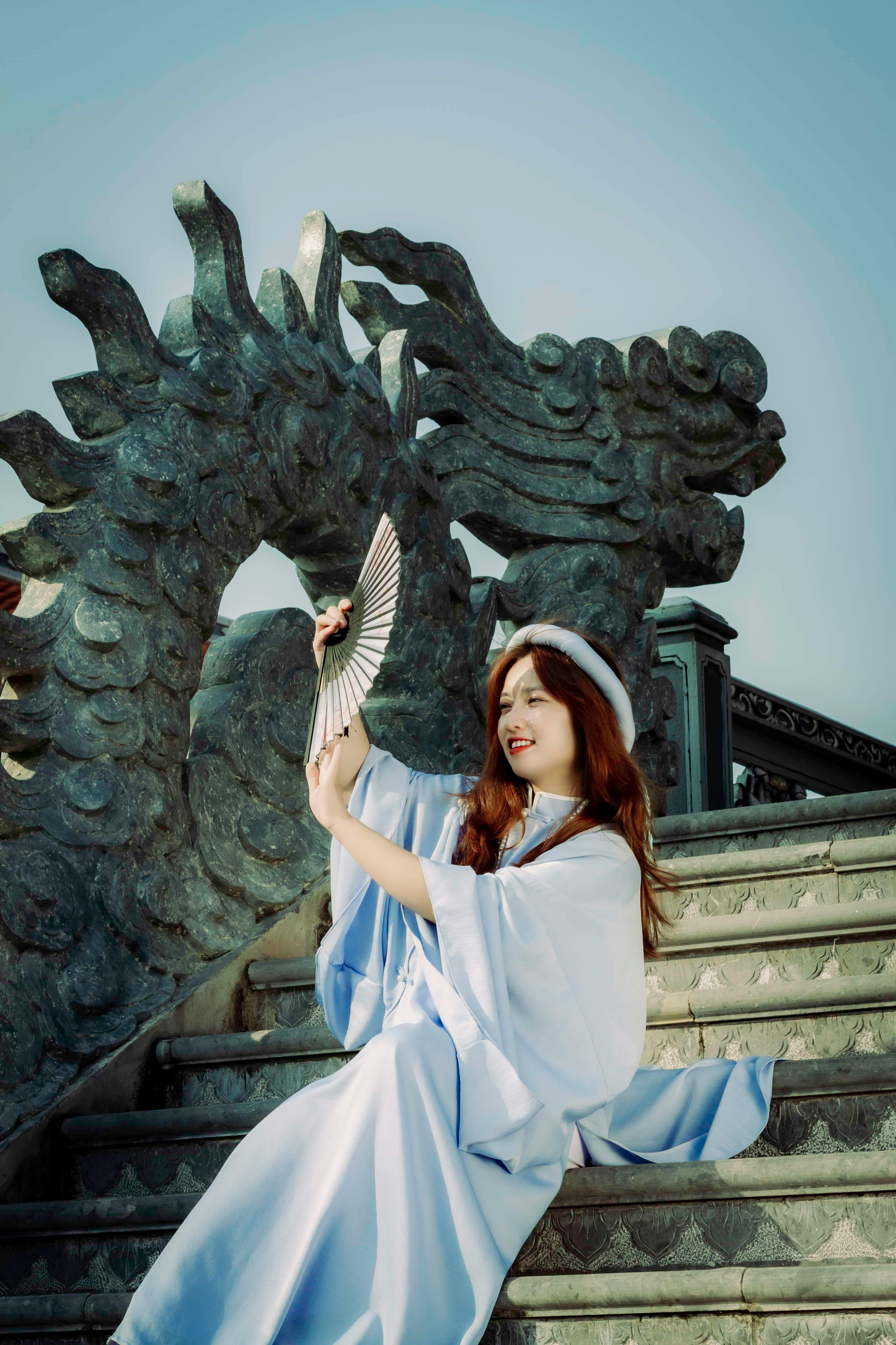 Elegantly dressed woman with a fan on stone steps next to a sculpture.