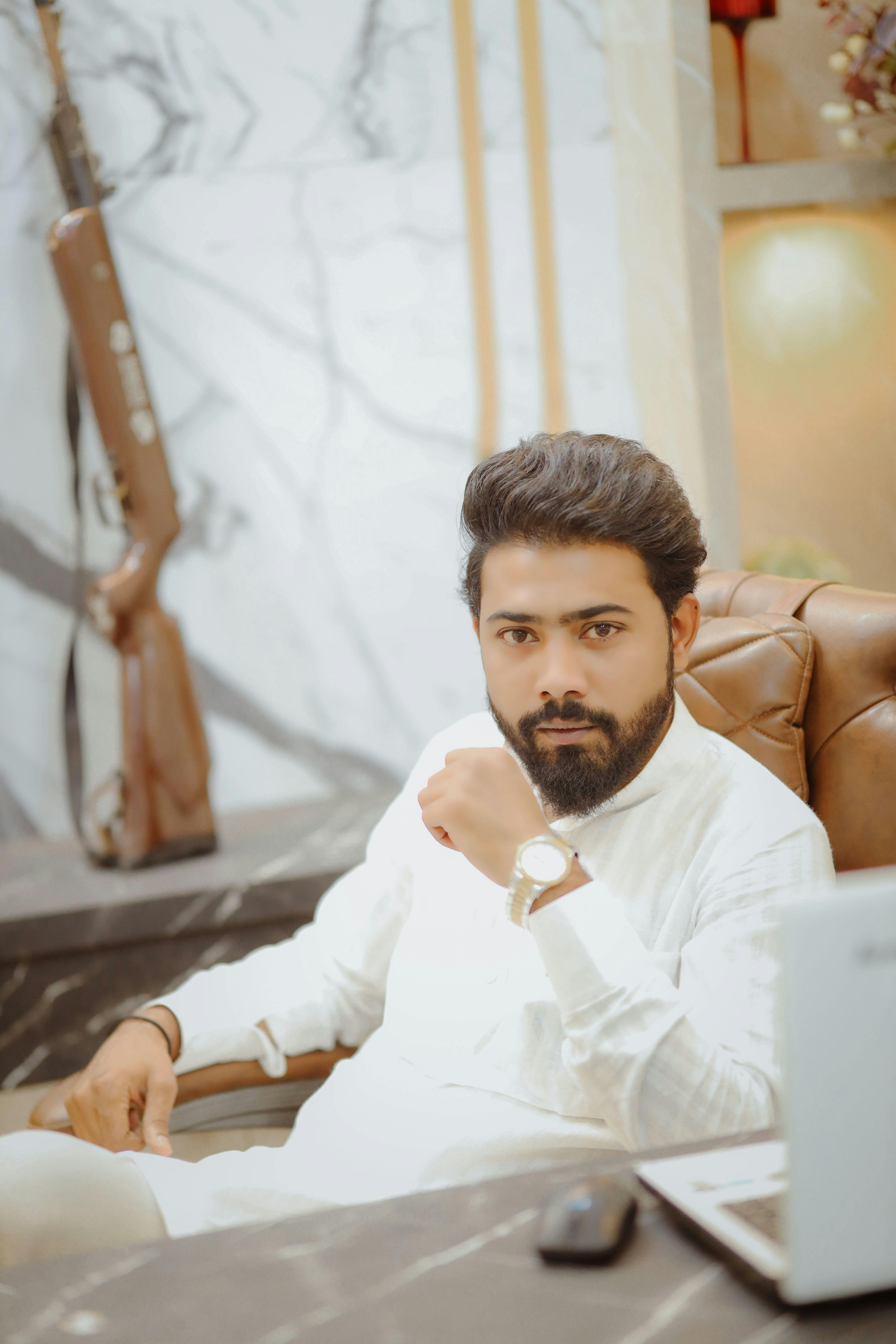 Fashionable bearded man in white attire posing confidently indoors by a table.