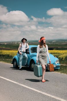 A stylish couple on a vintage road trip with a blue Volkswagen Beetle and suitcases.