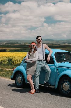 A young couple in retro attire posing by a vintage blue Volkswagen Beetle on a sunny day.