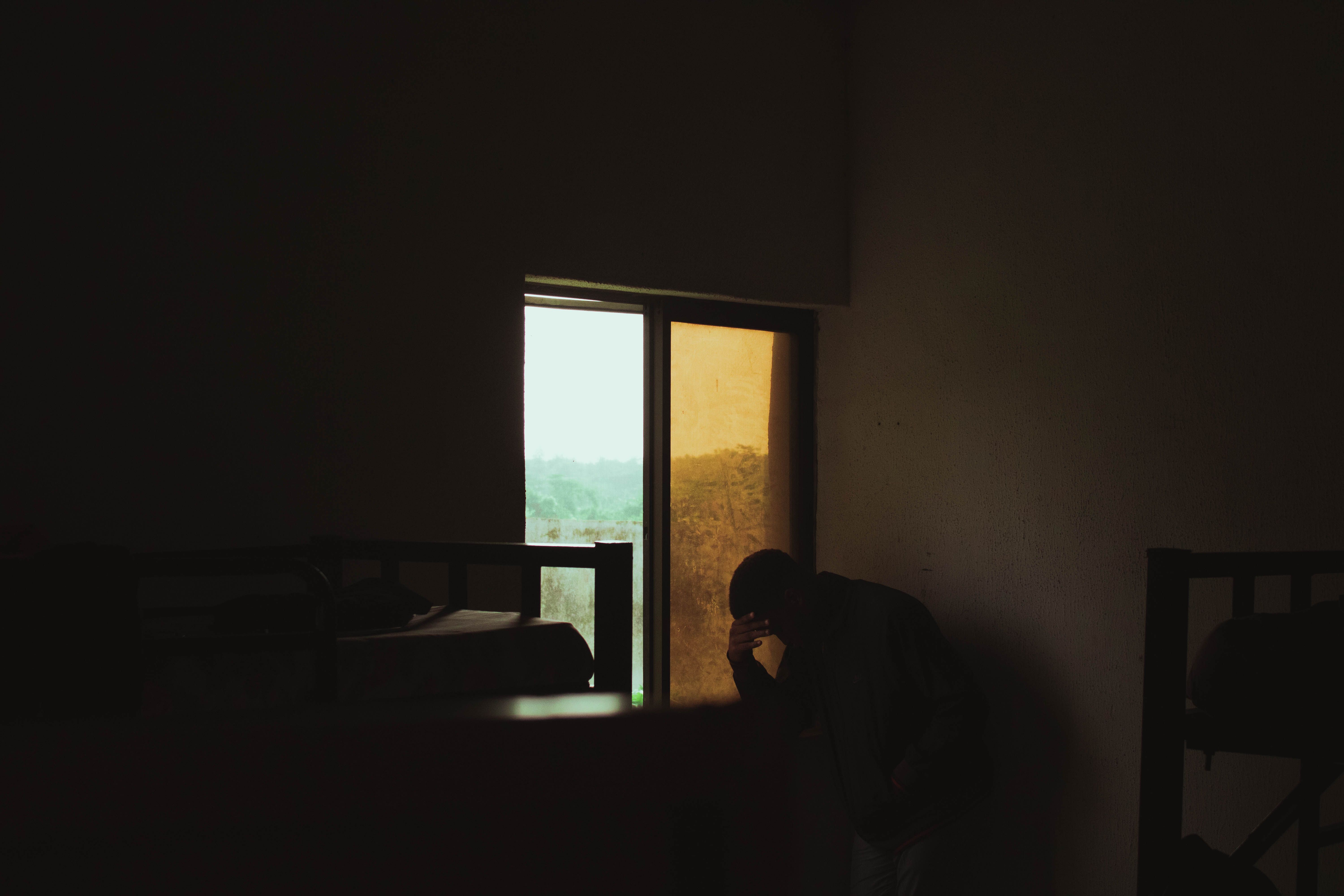 Silhouette of a man sitting alone in a dimly lit room by a window, evoking solitude.