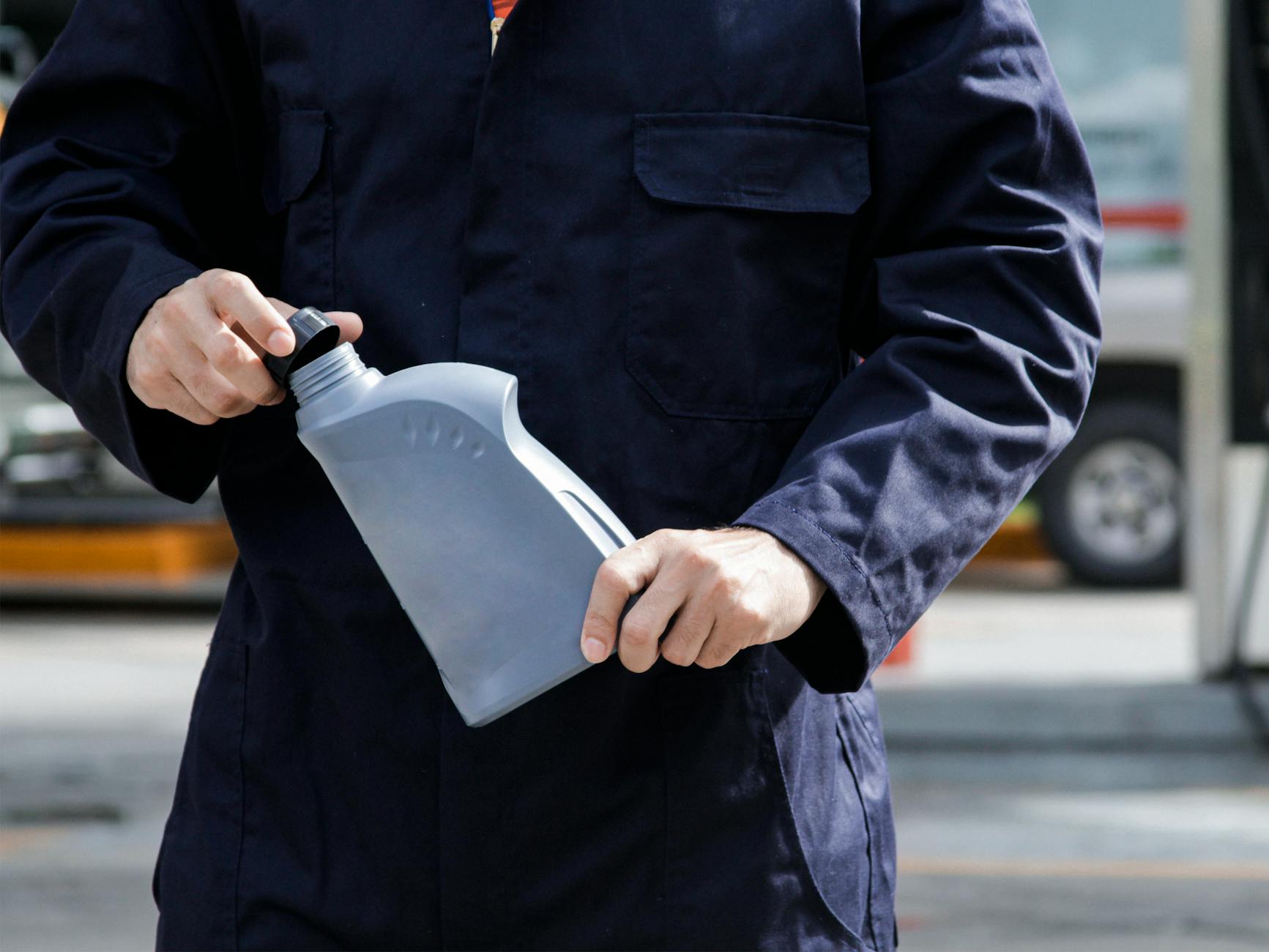 Close-up of a mechanic in a blue uniform holding a gray oil container on a city street.