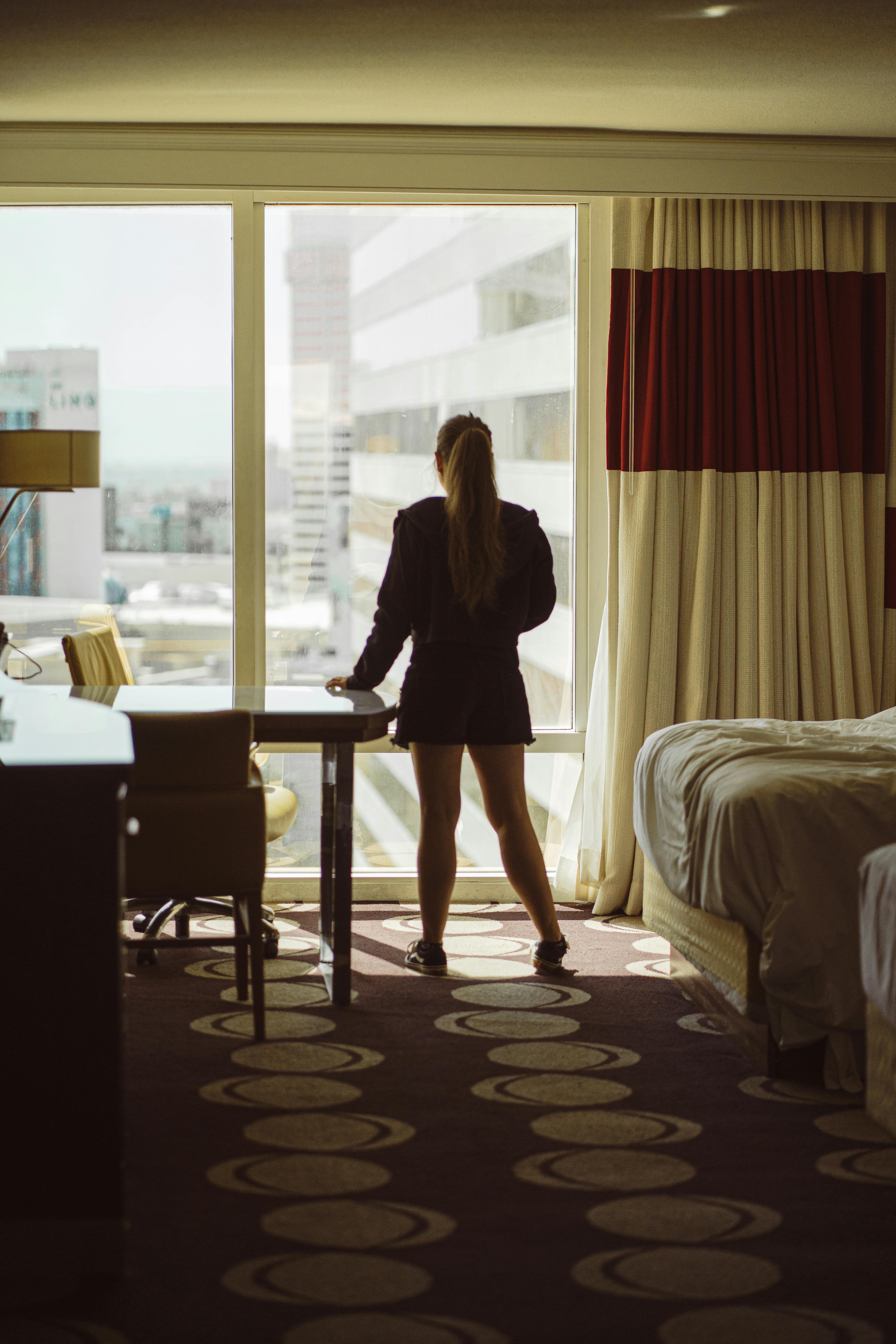 A woman stands by a hotel room window, viewing the Las Vegas skyline during the day.