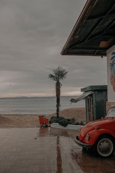 Vintage red car parked by a seaside view with palm in rainy weather, offering a nostalgic coastal vibe.
