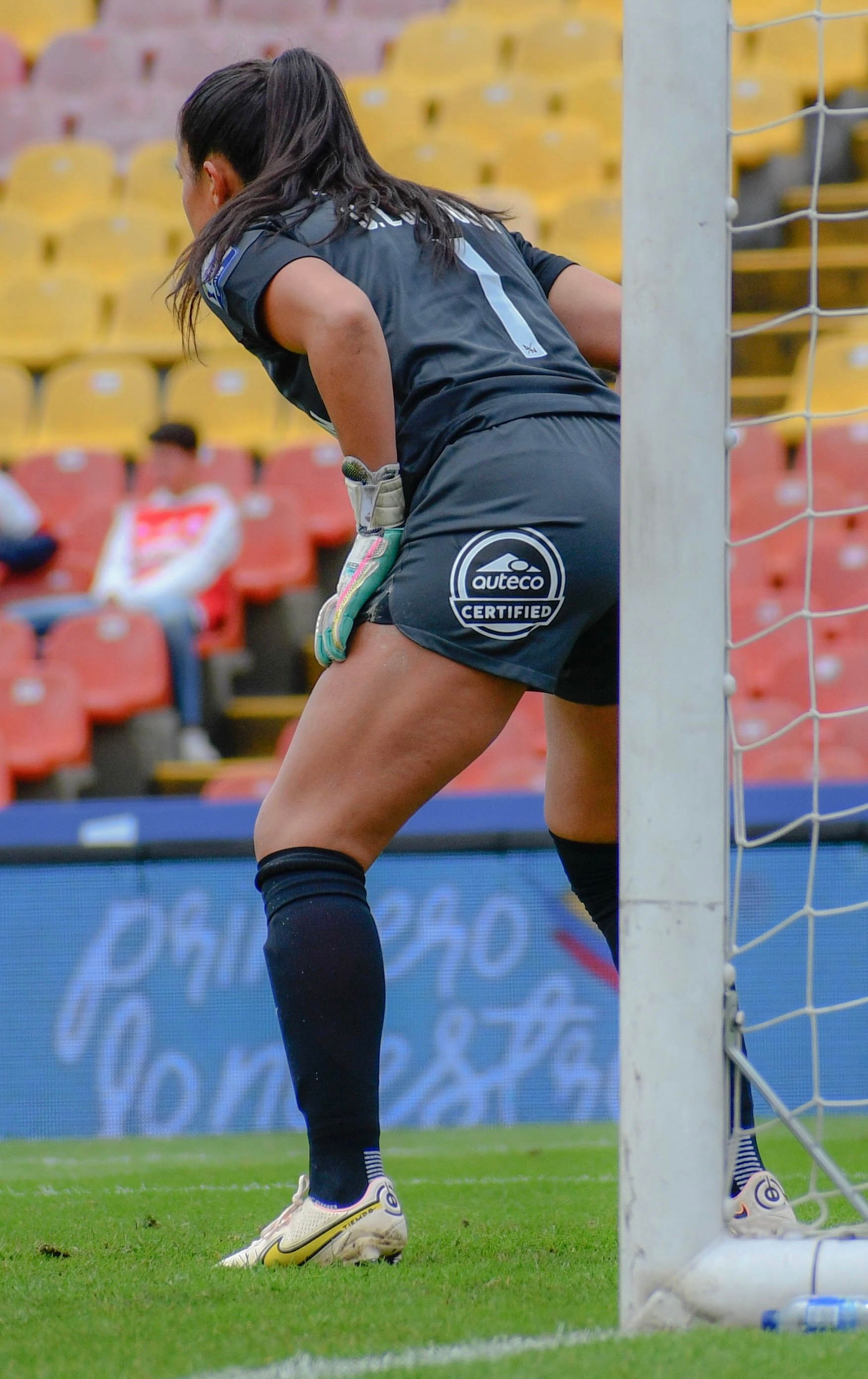 A female goalkeeper in action on a soccer field during a match, focusing near the goal post.