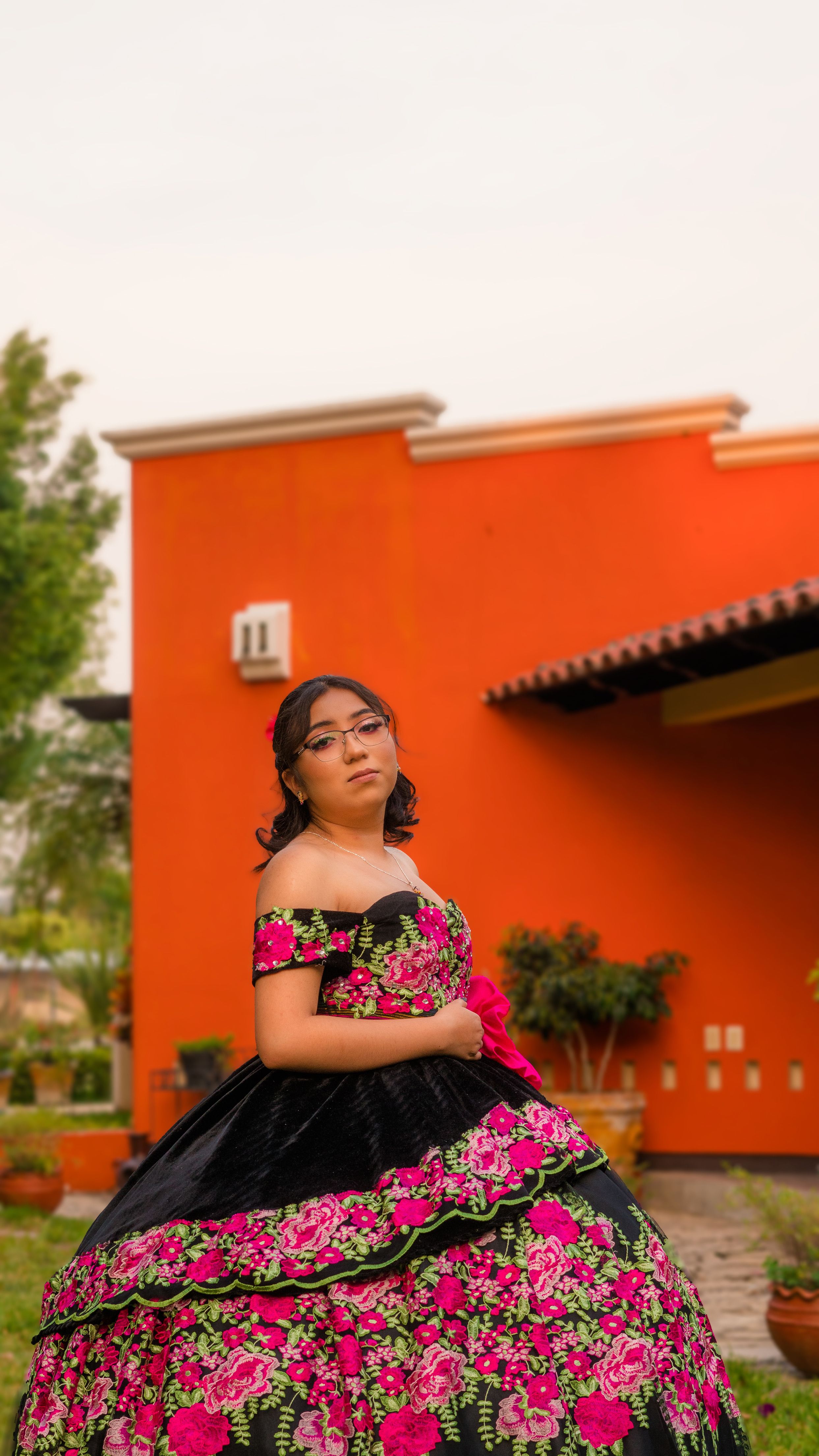 Woman in a detailed floral dress posing outdoors by an orange building wall.