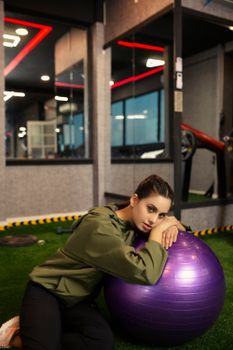 Woman in sportswear rests against a purple exercise ball in a modern gym setting.