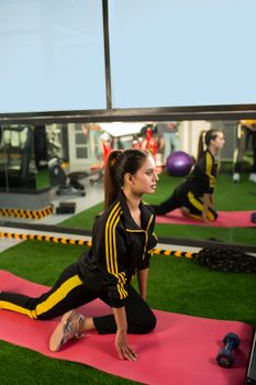 Woman in sportswear exercising in a gym, performing stretches on a mat next to a mirror.