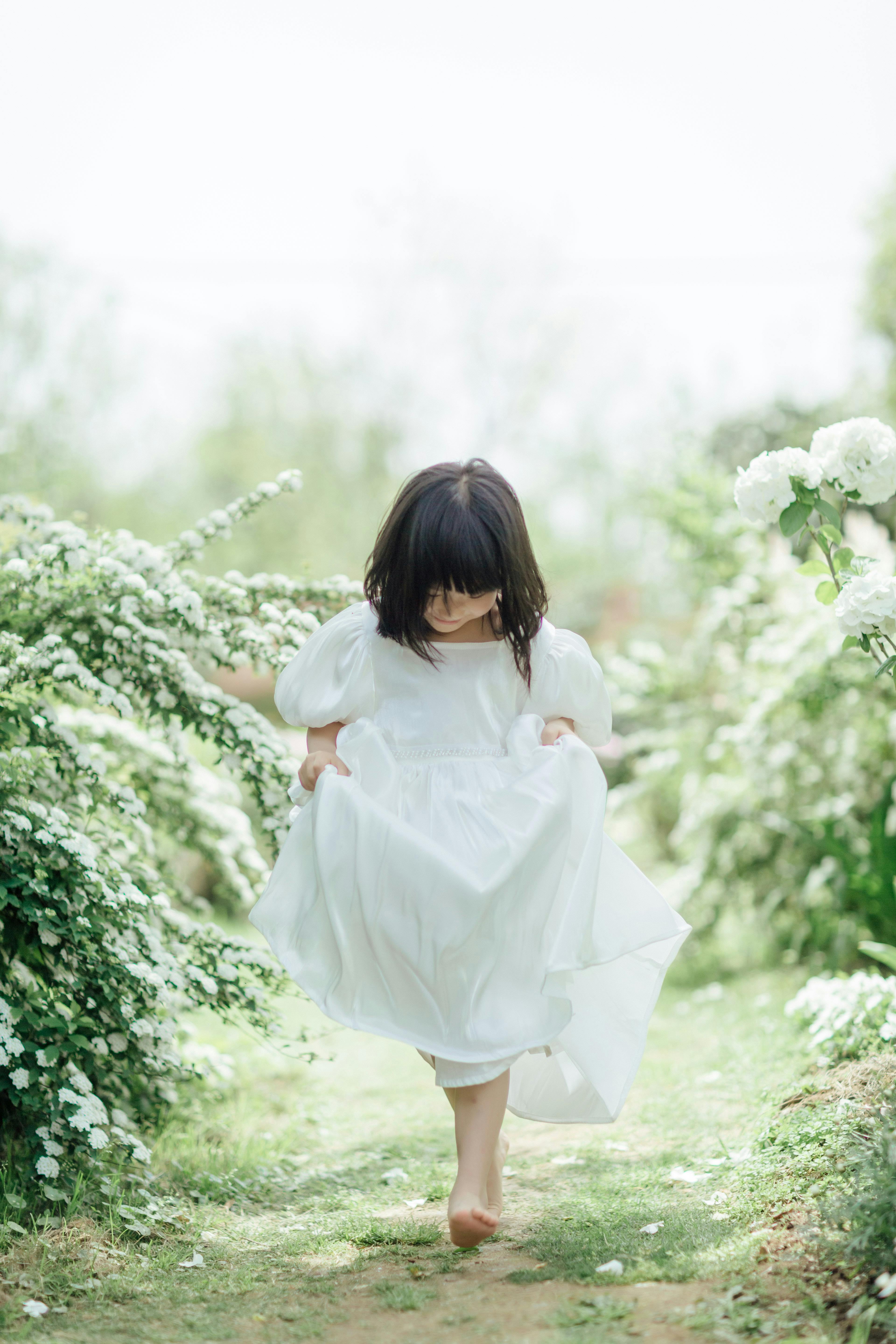 A young girl in a white dress walks barefoot among blooming shrubs in a serene garden setting.