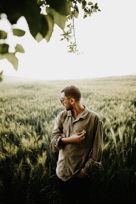Portrait of a stylish man with glasses in a lush green field, showcasing rural fashion.