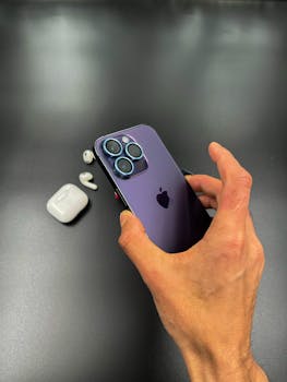 A close-up of an adult hand holding a smartphone with earbuds placed nearby on a black surface.