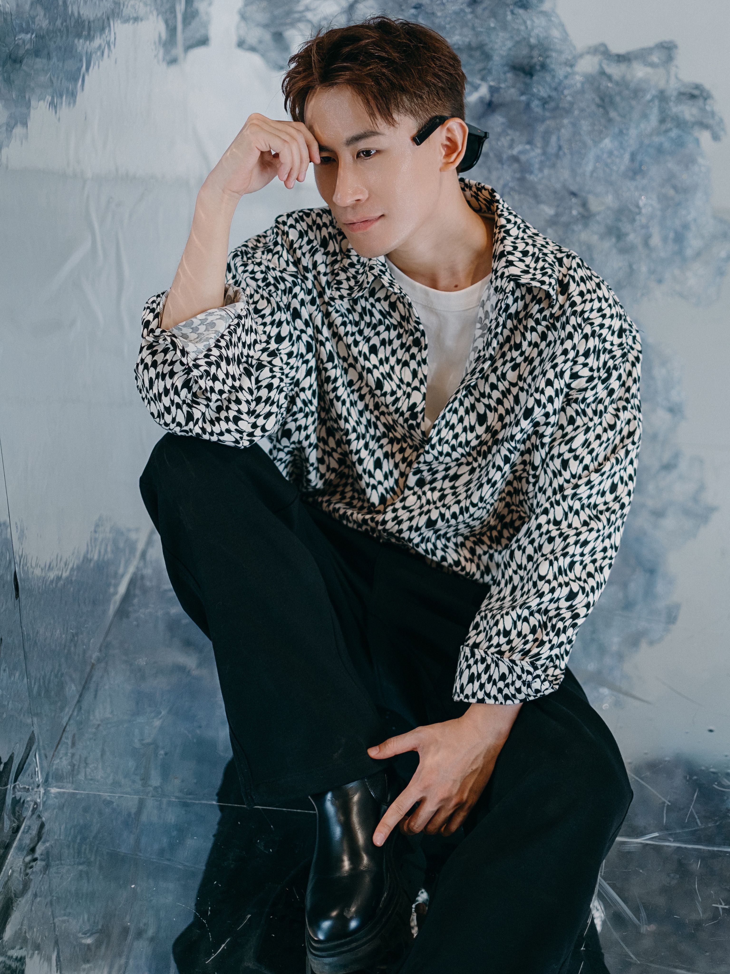 Portrait of a stylish man posing indoors with a contemplative expression, wearing a patterned shirt and black pants.