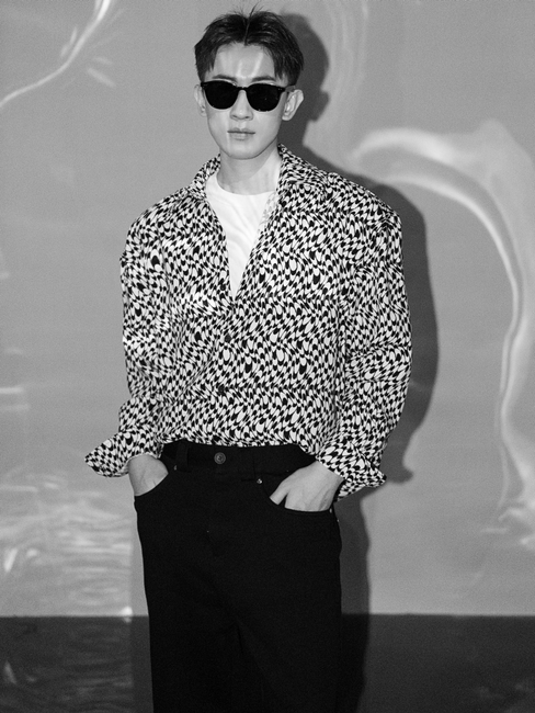 Black and white photo of a man with sunglasses and patterned shirt, hands in pockets.