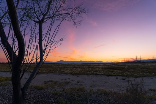 A tranquil desert landscape at sunset with silhouetted trees and distant mountains under a colorful sky.